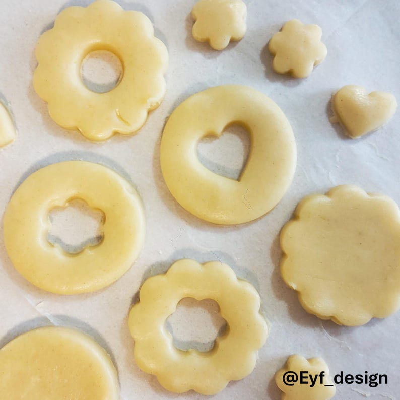 May include: Unbaked light beige cookie dough cut into various shapes including scalloped circles with center cutouts, a circle with a heart cutout, a solid heart, and small flower shapes. The text Eyf_design is visible in the bottom right corner.