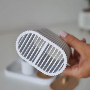 May include: A white and gray oval-shaped object with a ridged design and a series of vertical openings. The object is held in a person's hand. The background includes a white tray and a wooden soap dish.