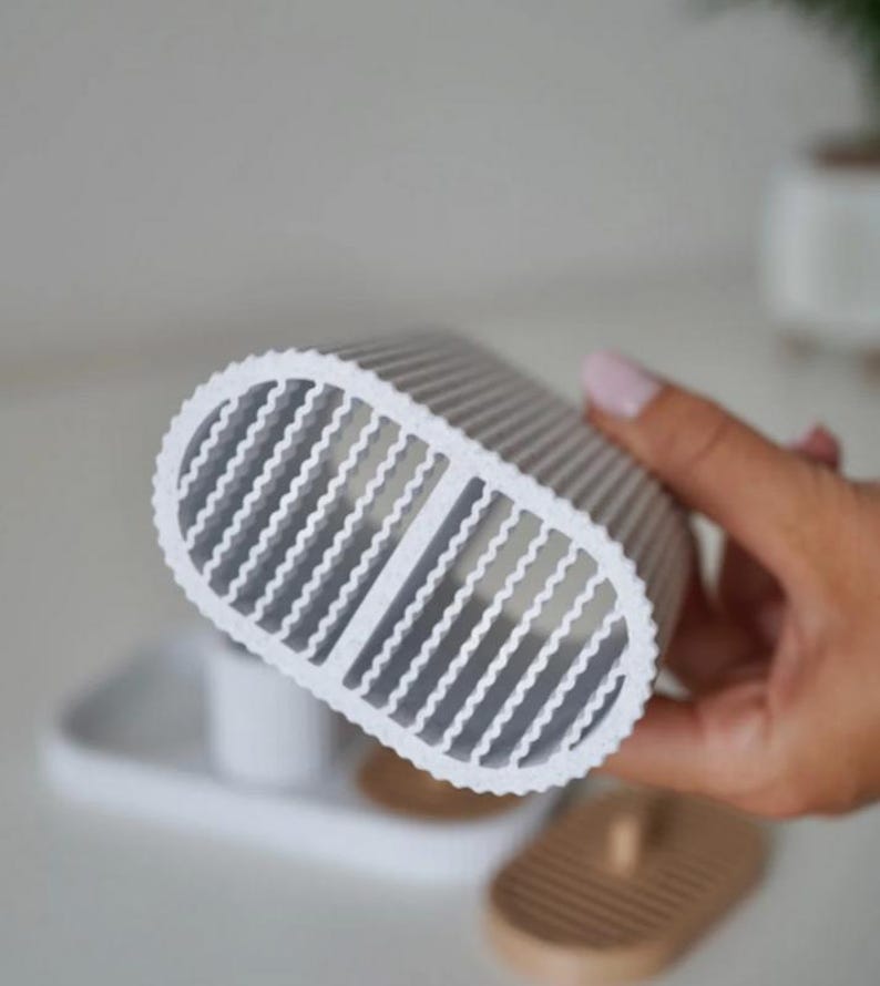 May include: A white and gray oval-shaped object with a ridged design and a series of vertical openings. The object is held in a person's hand. The background includes a white tray and a wooden soap dish.