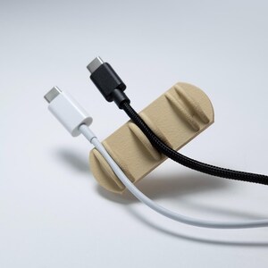 May include: A beige cable organiser with a rounded rectangular shape and three vertical slots. A white USB-C cable is secured in the first slot, and a black braided USB-C cable is positioned in the second slot, leaving the third slot empty.