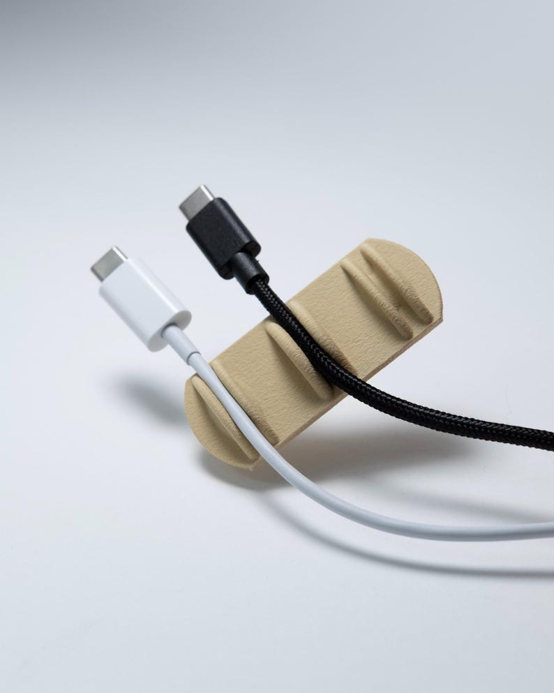 May include: A beige cable organiser with a rounded rectangular shape and three vertical slots. A white USB-C cable is secured in the first slot, and a black braided USB-C cable is positioned in the second slot, leaving the third slot empty.