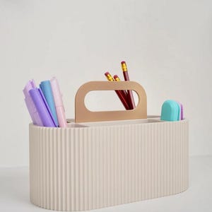 May include: A beige, ribbed desk organizer with a wooden handle. The organizer is filled with pencils, pens, and erasers in various colors. The design is simple and modern, suitable for office or home use.