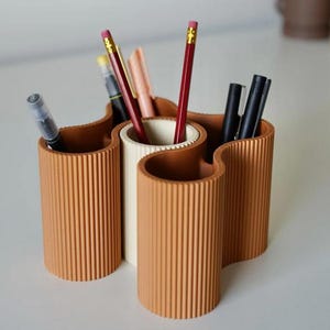 May include: A terracotta-colored desk organizer with a wavy design, holding pens and pencils. The organizer has three sections, with a central white section and two terracotta sections. The pencils are red and yellow.
