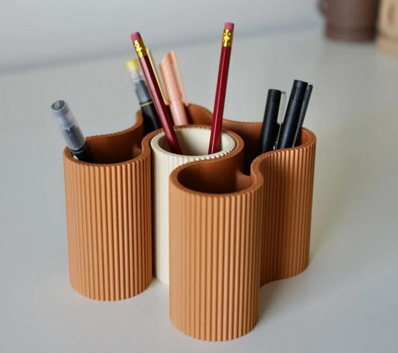 May include: A terracotta-colored desk organizer with a wavy design, holding pens and pencils. The organizer has three sections, with a central white section and two terracotta sections. The pencils are red and yellow.
