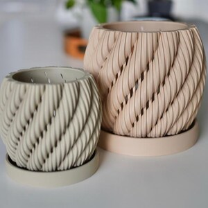 May include: Two decorative beige and off-white planters with a unique twisted rope design. The planters have matching saucers and are suitable for indoor or outdoor use. The larger planter is approximately 7 inches tall and 7 inches wide.