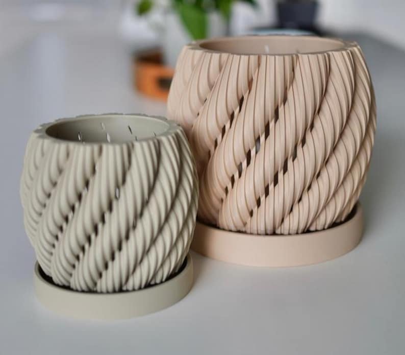 May include: Two decorative beige and off-white planters with a unique twisted rope design. The planters have matching saucers and are suitable for indoor or outdoor use. The larger planter is approximately 7 inches tall and 7 inches wide.