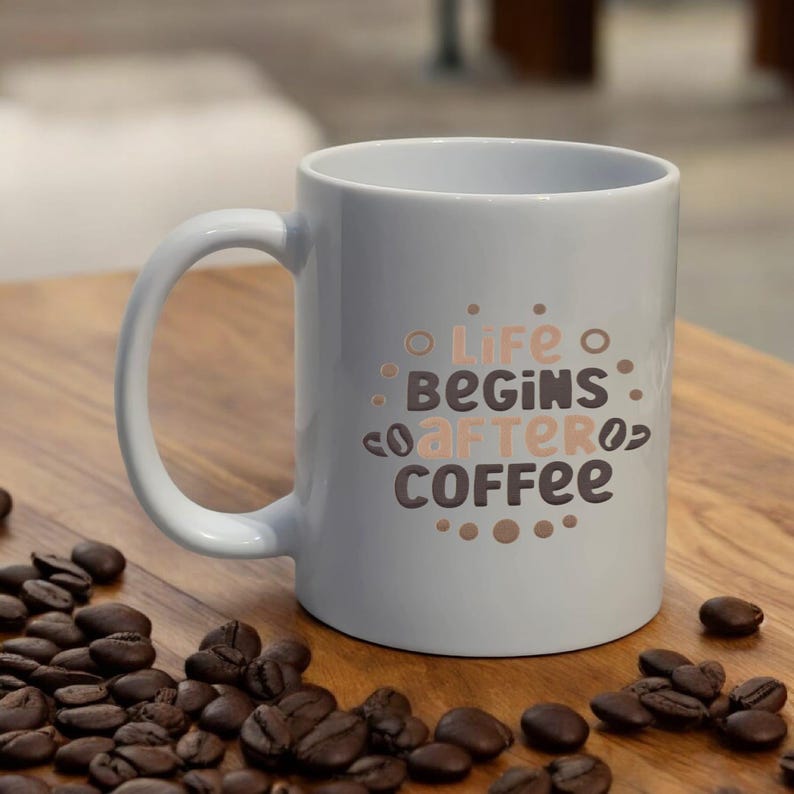 Puede incluir: Una taza de caf&eacute; de cer&aacute;mica blanca con la frase "Life Begins After Coffee" impresa en letras marrones y color melocot&oacute;n. La taza est&aacute; sobre una superficie de madera con granos de caf&eacute; esparcidos.