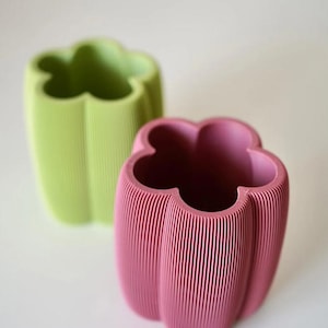 May include: Two decorative vases shaped like bell peppers. One is light green, the other is pink. Both have a ribbed texture and a four-lobed opening. The vases are set against a white background.
