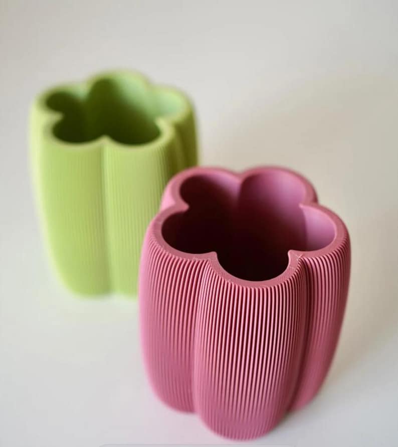 May include: Two decorative vases shaped like bell peppers. One is light green, the other is pink. Both have a ribbed texture and a four-lobed opening. The vases are set against a white background.