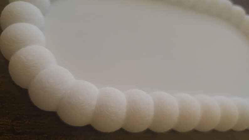 May include: Close-up of a white, round decorative object with a textured, beaded edge. The beads are spherical and evenly spaced, creating a continuous, elegant border. The background is a soft, off-white color.