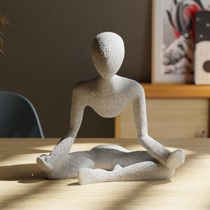 May include: A light gray, textured sculpture of a person in a seated yoga pose. The figure has a smooth, rounded head and arms extended to the sides. The sculpture is on a light wood surface.