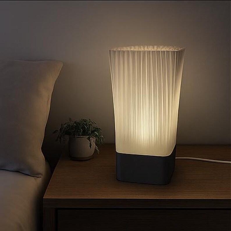 May include: A white, pleated lampshade sits atop a dark gray base, illuminated from within. The lamp is on a wooden nightstand next to a small potted plant and a bed with a pillow. A white cord extends from the lamp.