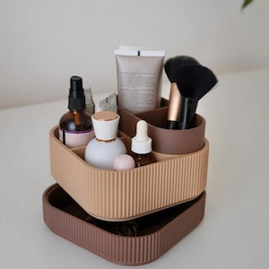 Op de afbeelding: Een tweekleurige, beige en bruine cosmetica-organizer met verticale ribbels. De bovenste laag bevat diverse schoonheidsproducten, waaronder flesjes, tubes en make-up borstels. De onderste laag is een effen bruin dienblad.