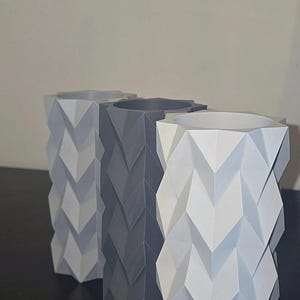Puede incluir: Tres jarrones geom&eacute;tricos en tonos grises y blancos. Los jarrones tienen un patr&oacute;n en zigzag y una parte superior cuadrada. Los jarrones est&aacute;n dispuestos en fila, con los colores cambiando gradualmente de claro a oscuro.