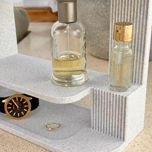 May include: A light gray speckled tiered organizer with vertical ribbed side detailing. The top tier holds a clear glass perfume bottle with a metallic cap and a smaller bottle with a gold lid. The bottom tier holds a black wristwatch and a gold ring.