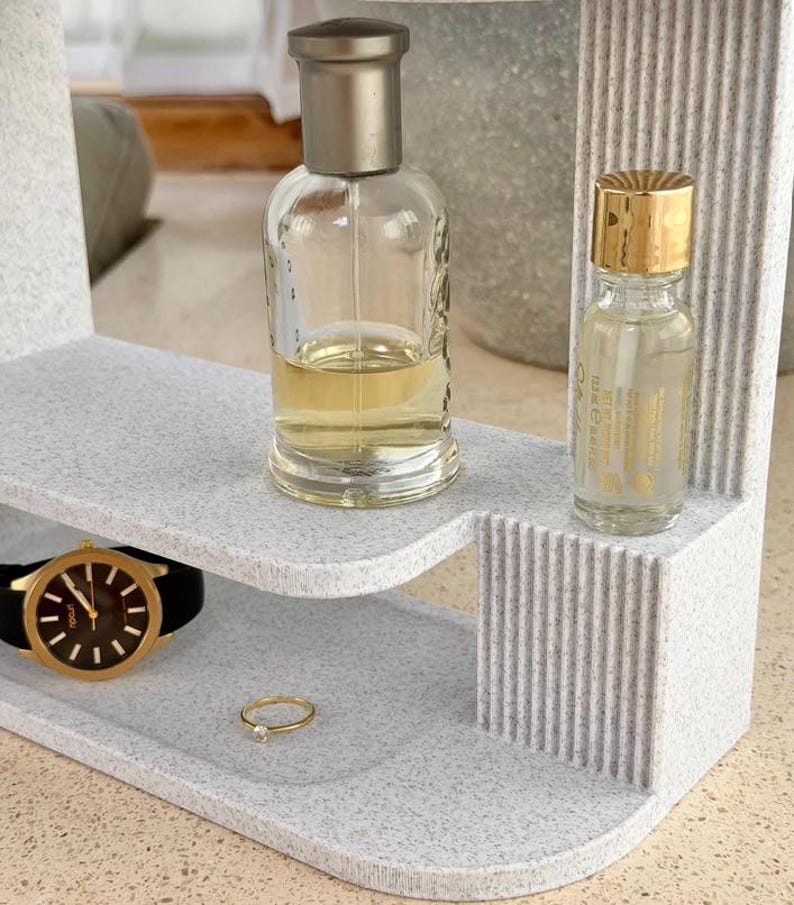 May include: A light gray speckled tiered organizer with vertical ribbed side detailing. The top tier holds a clear glass perfume bottle with a metallic cap and a smaller bottle with a gold lid. The bottom tier holds a black wristwatch and a gold ring.