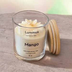 May include: A clear glass candle jar with a white candle and a wooden lid. The candle is labeled "Lummus Scented candles" and "Mango". The candle is topped with a white flower. The label also states "100% Natural soy wax Handmade candle 8 oz".