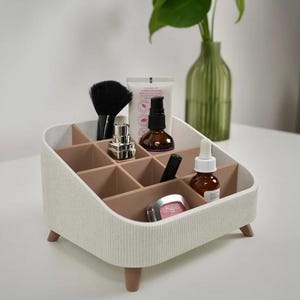 Puede incluir: Un organizador de maquillaje blanco y beige con m&uacute;ltiples compartimentos, que contiene varios productos de belleza. El organizador tiene un exterior texturizado y se apoya en peque&ntilde;as patas de madera. Un pincel de maquillaje negro y otros art&iacute;culos cosm&eacute;ticos son visibles.