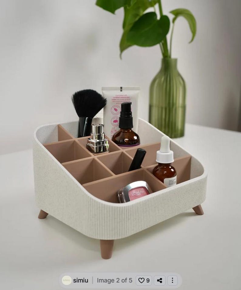 Puede incluir: Un organizador de maquillaje blanco y beige con m&uacute;ltiples compartimentos, que contiene varios productos de belleza. El organizador tiene un exterior texturizado y se apoya en peque&ntilde;as patas de madera. Un pincel de maquillaje negro y otros art&iacute;culos cosm&eacute;ticos son visibles.