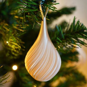 May include: A pear-shaped Christmas ornament with a smooth, pearlescent off-white finish and a distinct spiraling, ribbed texture that tapers toward the top. The ornament features a thin, metallic silver loop for hanging from a tree branch.