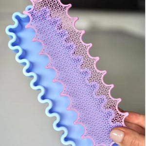 May include: A light blue, wavy-edged tray with a pink, patterned, decorative overlay. The overlay has a repeating geometric design and a scalloped edge. The tray is held by a person.