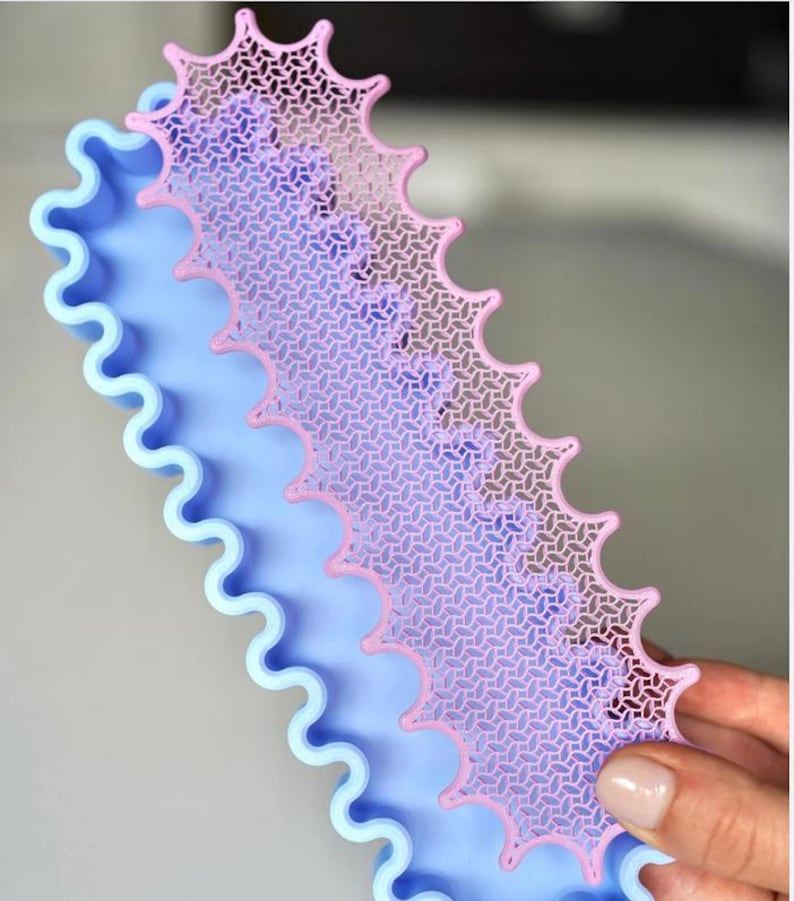 May include: A light blue, wavy-edged tray with a pink, patterned, decorative overlay. The overlay has a repeating geometric design and a scalloped edge. The tray is held by a person.