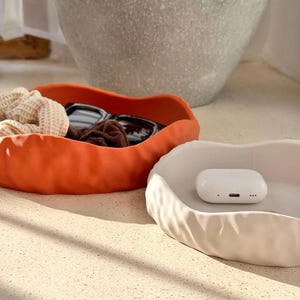 May include: Two shallow, wavy-edged bowls, one in matte orange and one in matte white. The orange bowl contains a beige textured scrunchie and black sunglasses. The white bowl holds a small, white, rectangular wireless earbud charging case.