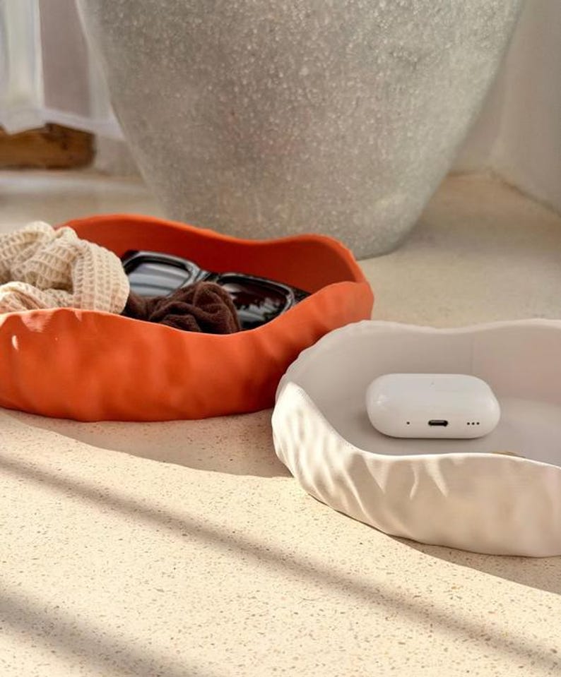 May include: Two shallow, wavy-edged bowls, one in matte orange and one in matte white. The orange bowl contains a beige textured scrunchie and black sunglasses. The white bowl holds a small, white, rectangular wireless earbud charging case.