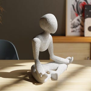May include: A light gray stone sculpture of a person in a seated yoga pose. The figure has a smooth, rounded head and body, with arms and legs in a meditative position. The sculpture is on a light wood surface.