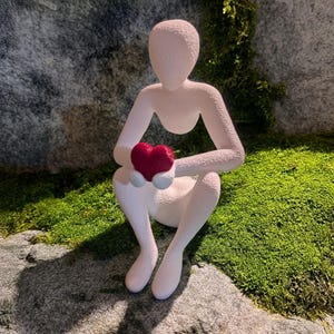 Puede incluir: Una figura blanca de una persona sentada sosteniendo un coraz&oacute;n rojo. La figura est&aacute; colocada sobre una superficie de piedra cubierta de musgo verde, con un fondo de roca gris texturizada.