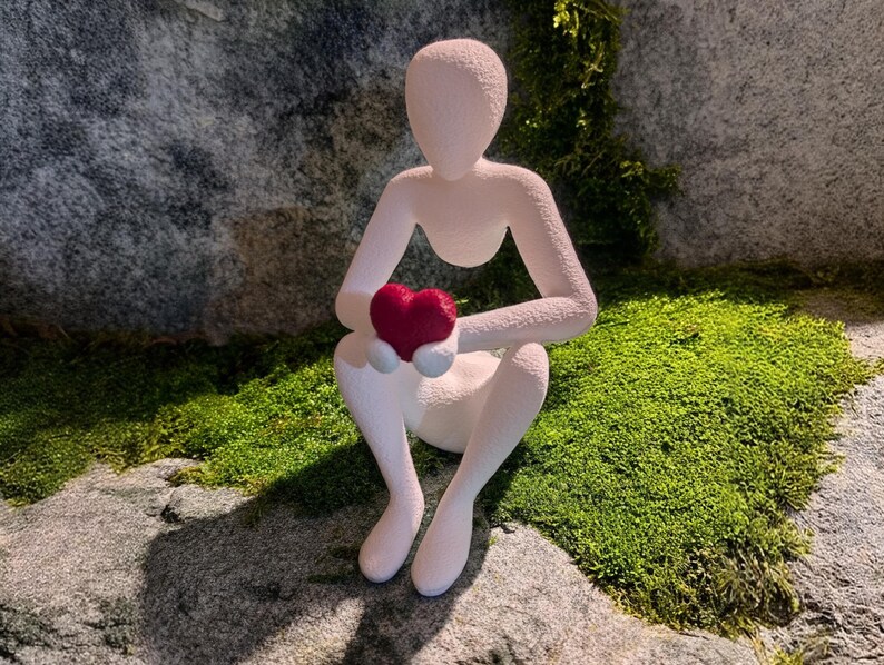 Puede incluir: Una figura blanca de una persona sentada sosteniendo un coraz&oacute;n rojo. La figura est&aacute; colocada sobre una superficie de piedra cubierta de musgo verde, con un fondo de roca gris texturizada.