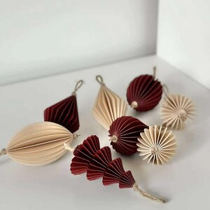 May include: An assortment of decorative paper ornaments in shades of burgundy and cream. The ornaments include geometric shapes, spheres, and a stylized leaf design, all with textured, folded paper construction. Each ornament has a string for hanging.