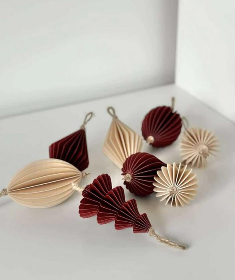 May include: An assortment of decorative paper ornaments in shades of burgundy and cream. The ornaments include geometric shapes, spheres, and a stylized leaf design, all with textured, folded paper construction. Each ornament has a string for hanging.