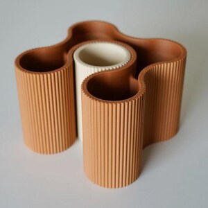 May include: A terracotta-colored, wavy vase with a central white cylinder. The vase has a ribbed texture and a unique, organic shape. The design features multiple openings, perfect for displaying flowers or other decorative items.