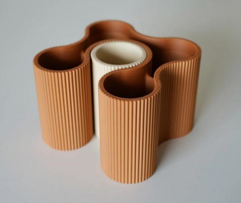 May include: A terracotta-colored, wavy vase with a central white cylinder. The vase has a ribbed texture and a unique, organic shape. The design features multiple openings, perfect for displaying flowers or other decorative items.