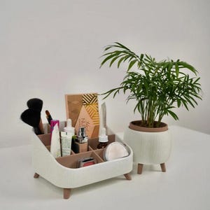Puede incluir: Un organizador de maquillaje blanco y marr&oacute;n con varios productos de belleza y brochas. Una planta en maceta con hojas verdes est&aacute; a la derecha. El organizador tiene varios compartimentos y est&aacute; sobre patas de madera.