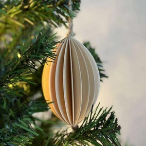 May include: A cream-colored, paper Christmas ornament in an oval shape, suspended from a twine string. The ornament has a pleated design, with vertical lines creating a textured appearance. The ornament hangs on a green Christmas tree branch.
