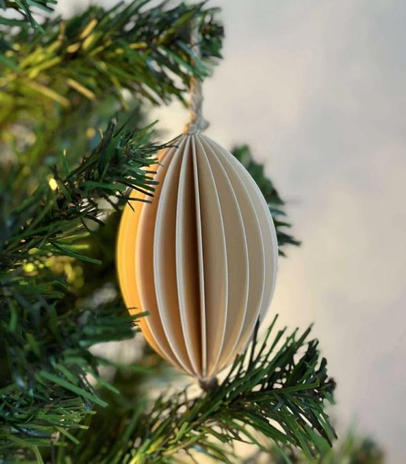 May include: A cream-colored, paper Christmas ornament in an oval shape, suspended from a twine string. The ornament has a pleated design, with vertical lines creating a textured appearance. The ornament hangs on a green Christmas tree branch.
