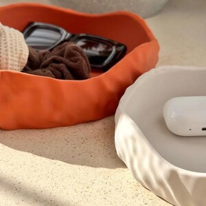 May include: Two irregular, wavy-edged trinket dishes, one in matte burnt orange and one in matte off-white. The orange dish contains black sunglasses and a brown scrunchie, while the white dish holds a small, smooth white rectangular case.