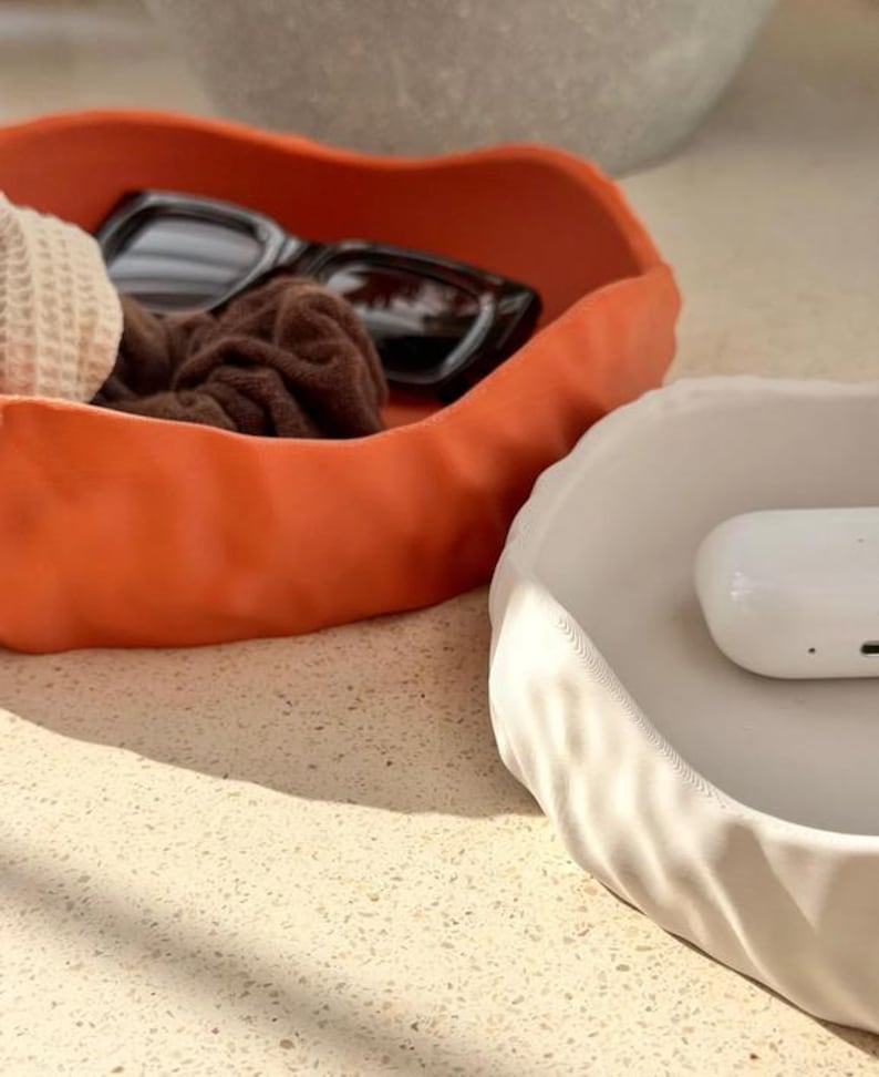 May include: Two irregular, wavy-edged trinket dishes, one in matte burnt orange and one in matte off-white. The orange dish contains black sunglasses and a brown scrunchie, while the white dish holds a small, smooth white rectangular case.