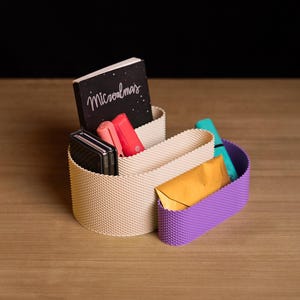 May include: A beige and purple desk organiser with a textured, grid-like surface. The organiser holds a black notebook with the text "Microalmas" on the cover, a coral-coloured pen, a black folding phone, a teal marker, and a gold envelope.