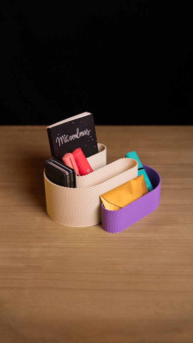May include: A beige and purple desk organiser with a textured, grid-like surface. The organiser holds a black notebook with the text "Microalmas" on the cover, a coral-coloured pen, a black folding phone, a teal marker, and a gold envelope.
