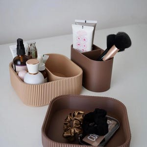 May include: A collection of neutral-toned organizers for cosmetics and accessories. The set includes a ribbed, divided tray, a divided cup for brushes and tubes, and a square tray with scrunchies and a compact. The containers are in shades of beige and brown.