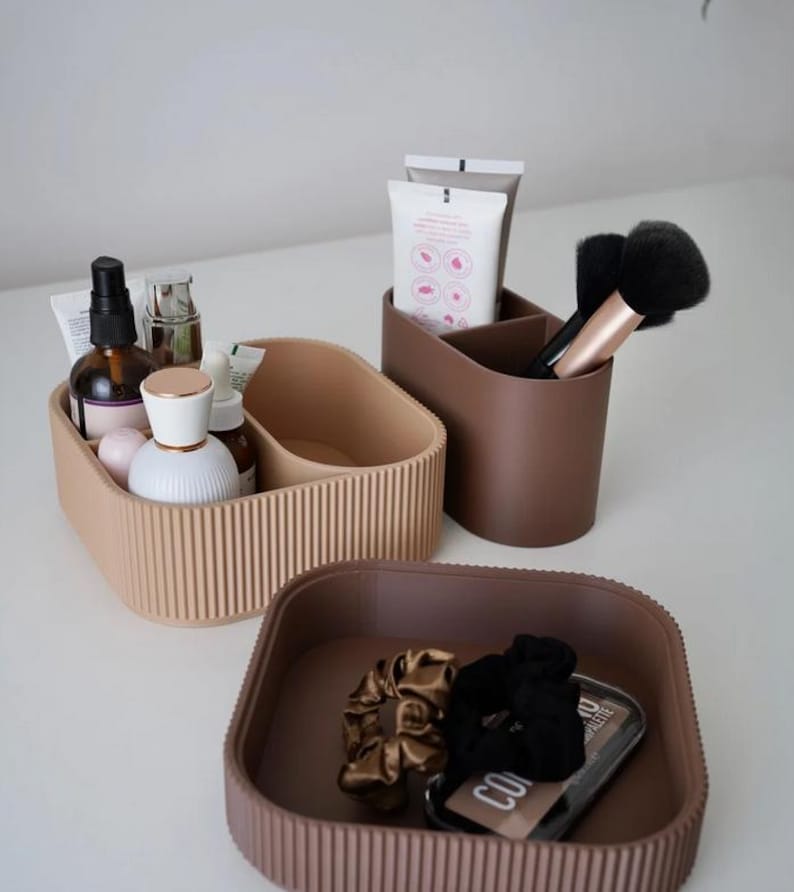 May include: A collection of neutral-toned organizers for cosmetics and accessories. The set includes a ribbed, divided tray, a divided cup for brushes and tubes, and a square tray with scrunchies and a compact. The containers are in shades of beige and brown.