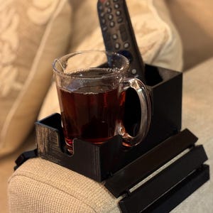 Puede incluir: Una bandeja negra para el brazo del sof&aacute; con un portavasos y un soporte para el mando a distancia incorporados. Una taza de cristal llena de un l&iacute;quido oscuro est&aacute; en el portavasos. Un mando a distancia negro descansa en el soporte. La bandeja est&aacute; sobre un brazo de sof&aacute; beige.