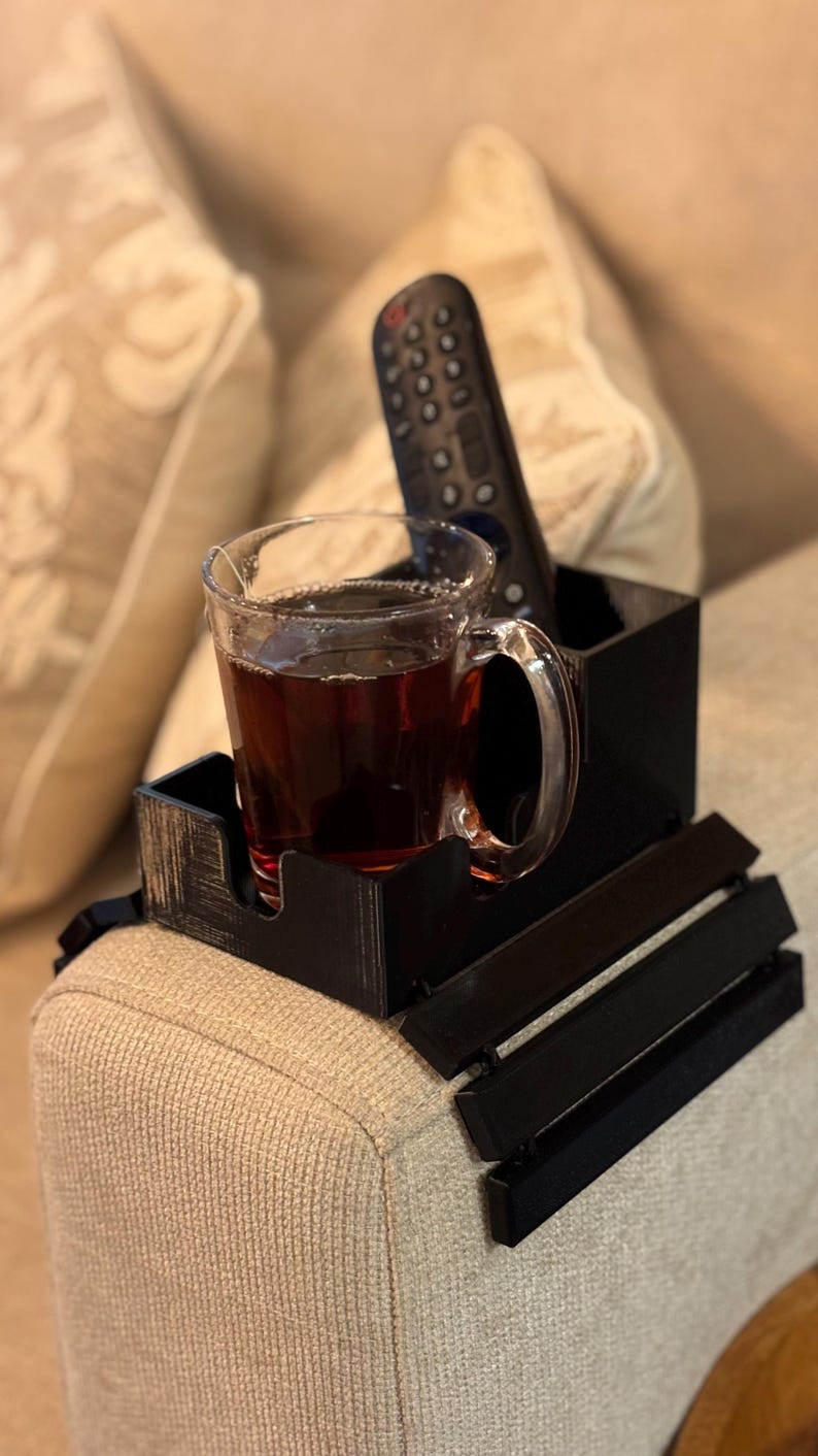 Puede incluir: Una bandeja negra para el brazo del sof&aacute; con un portavasos y un soporte para el mando a distancia incorporados. Una taza de cristal llena de un l&iacute;quido oscuro est&aacute; en el portavasos. Un mando a distancia negro descansa en el soporte. La bandeja est&aacute; sobre un brazo de sof&aacute; beige.