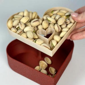 May include: A heart-shaped serving dish filled with pistachios, with a matching heart-shaped base. The top dish is beige, and the base is a textured, deep red. The pistachios are shelled and ready to eat.