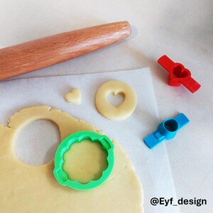May include: A wooden rolling pin, a green scalloped cookie cutter, a red heart-shaped plunger cutter, a blue plunger cutter, and raw cookie dough with a heart-shaped cutout and a small heart piece. The text @Eyf_design is visible in the corner.