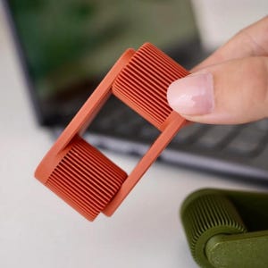 May include: A close-up of a coral-colored lint roller with a rectangular frame and a cylindrical rolling head. The roller is held in a person's hand, with a blurred background of a laptop and a second green roller.