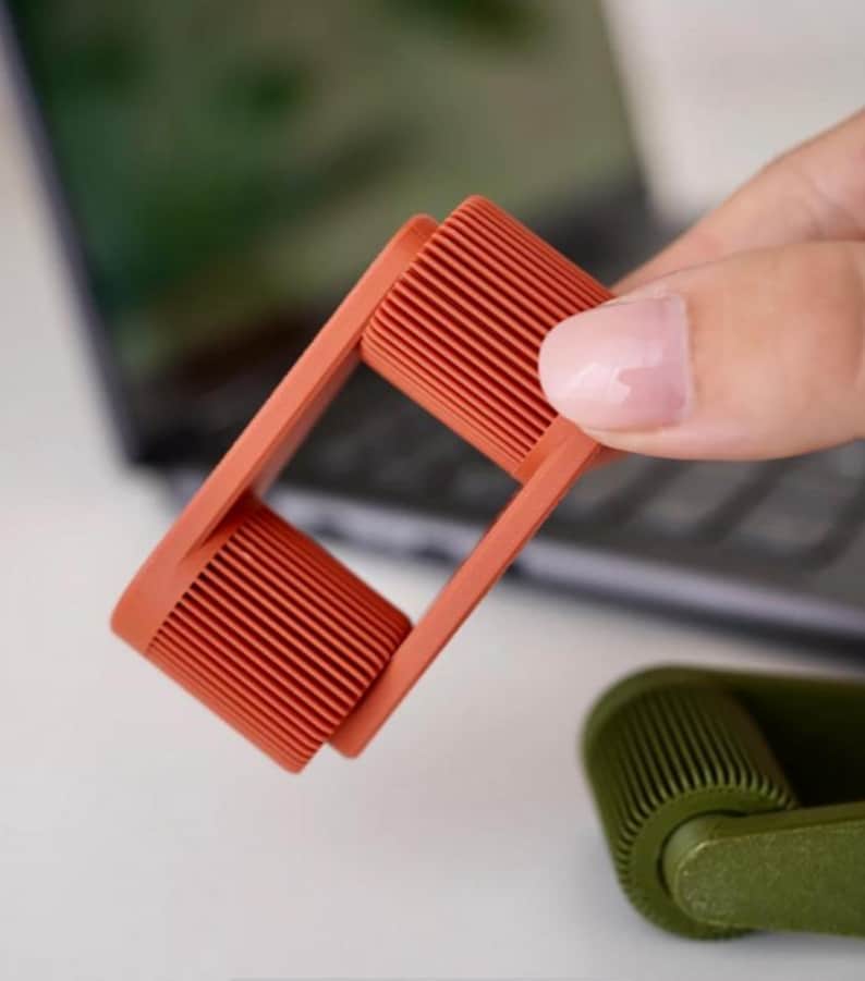May include: A close-up of a coral-colored lint roller with a rectangular frame and a cylindrical rolling head. The roller is held in a person's hand, with a blurred background of a laptop and a second green roller.